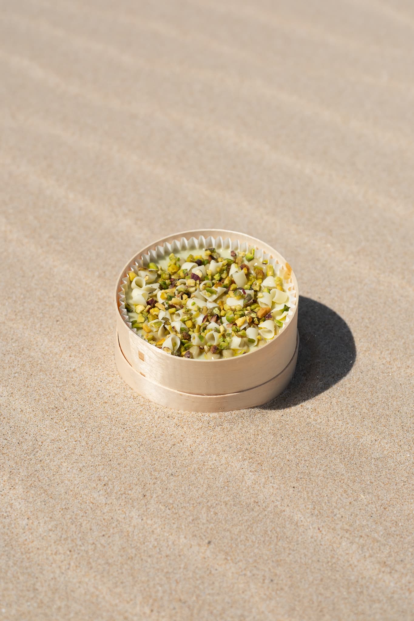 Cheesecake de pistacho cubierta de pistacho troceado, en su caja de madera sobre arena de playa.